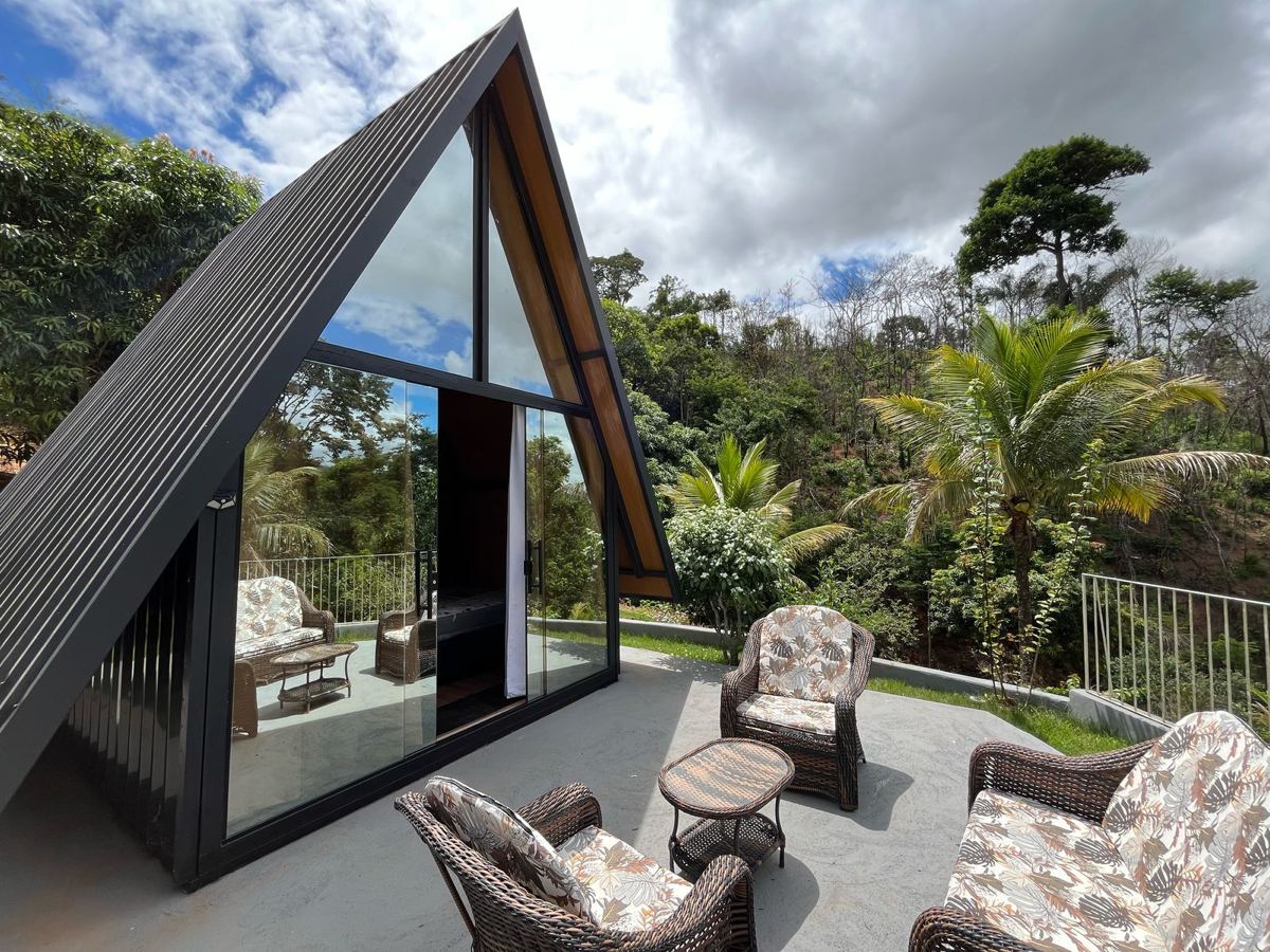 Casinha com fachada de vidro e terraço com poltronas, cercada de mata